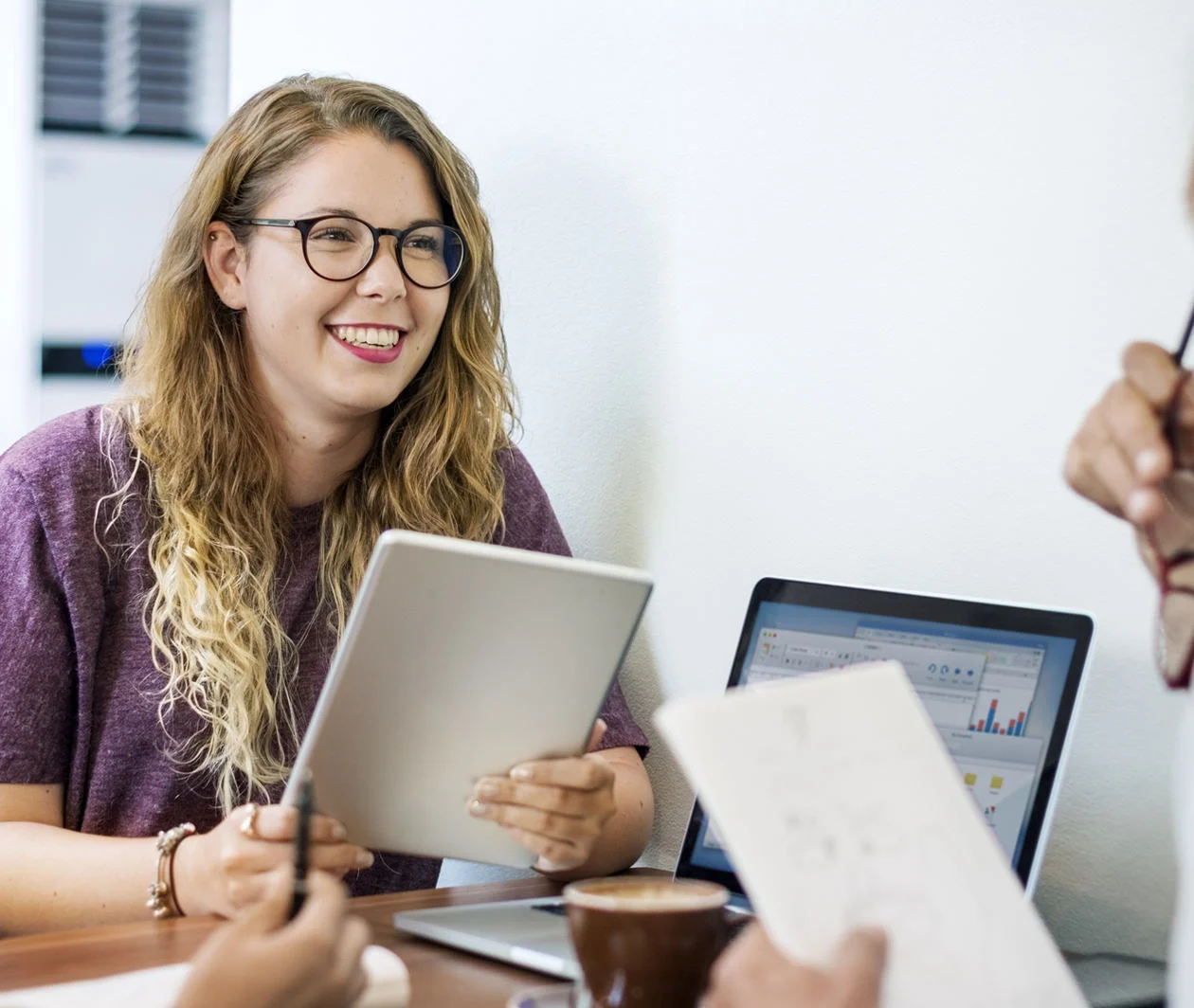 Three colleagues sit in a meeting around a table with a laptop on it displaying charts, a cup of coffee in a saucer. Each holds a pad of paper and a pen. The two people we see the faces of are happy.