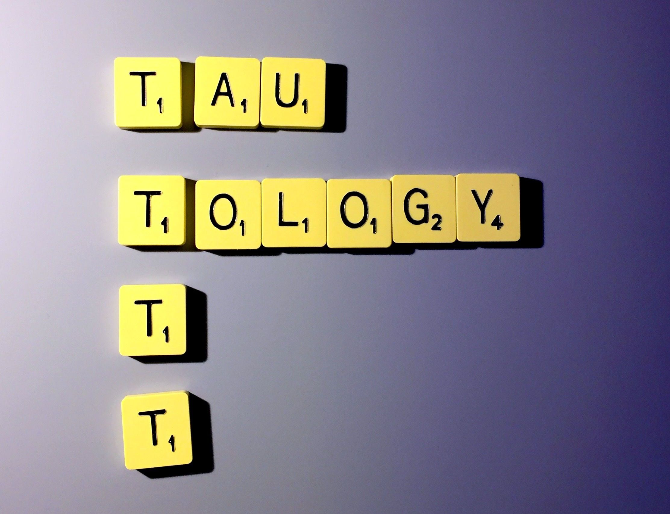 The word 'tautology' spelt out in letter tiles, split so that the tee of 'tau' is above the tee of 'tology', with two further tees underneath, so there is a column of 4 tees.