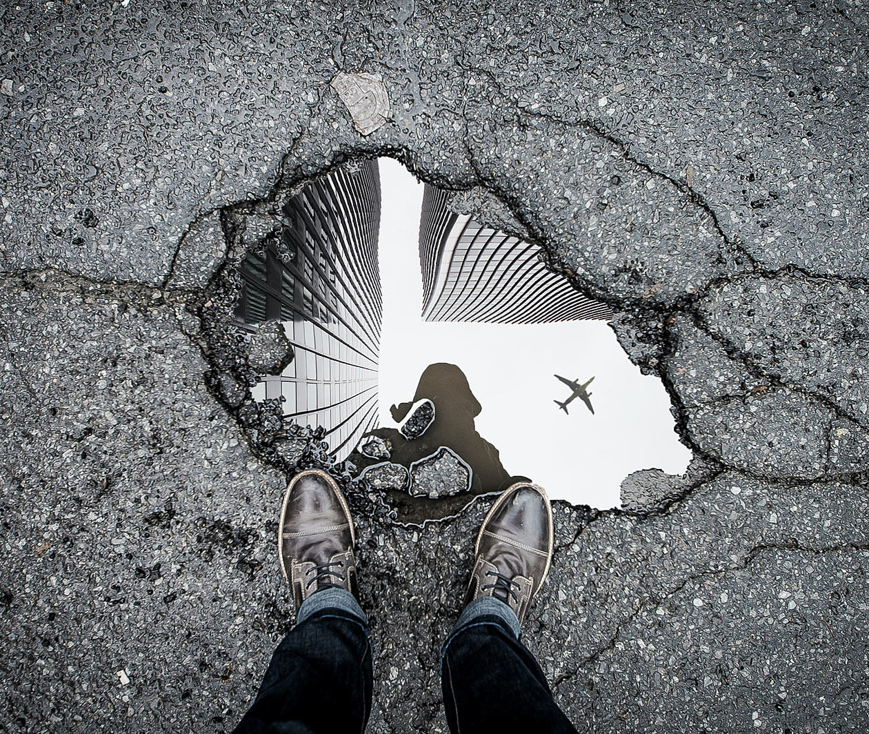 A view into a puddle in a pothole on the road with the reflection of a plane and skyscrapers in the water