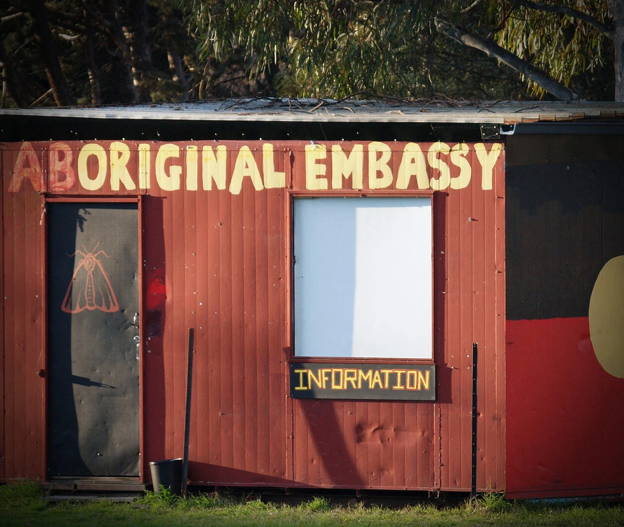 A small shed painted with the Australian Aboriginal flag on the side and with a sign above the door stating 'Aboriginal Embassy'