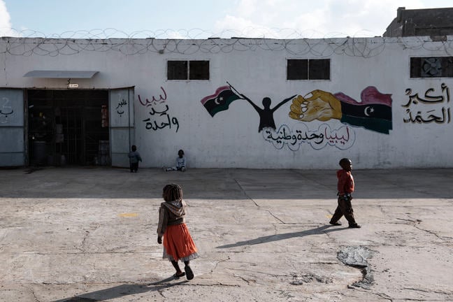 A very young girl in a bright orange skirt and a very young boy in a red shirt are outside a building with wire on the roof and graffiti on the white cement wall. The graphic is arabic writing with a silhouette of a person holding flags.