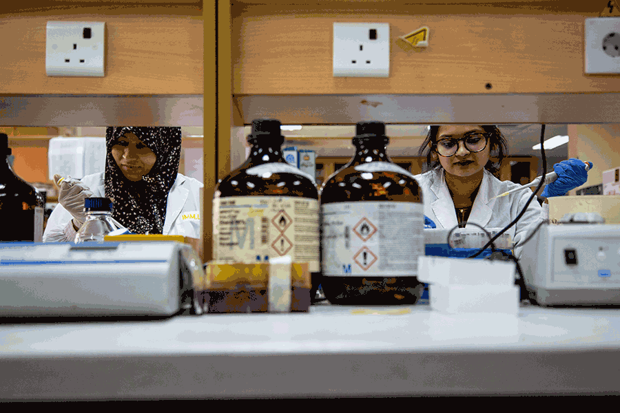 Two women wearing laboratory coats and gloves are standing behind a table with two big amber bottles and laboratory equipment. They are each holding a pipette to test samples for pathogens.