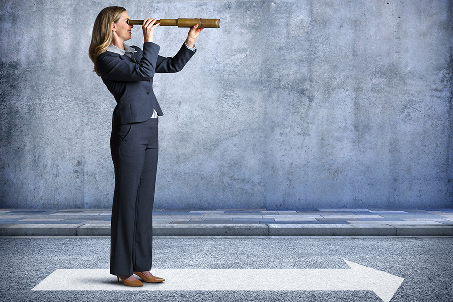 An arrow painted on the roadway points the way for a businesswoman as she stands at a profile to the camera and looks through a spyglass in the direction that the arrow is pointing.