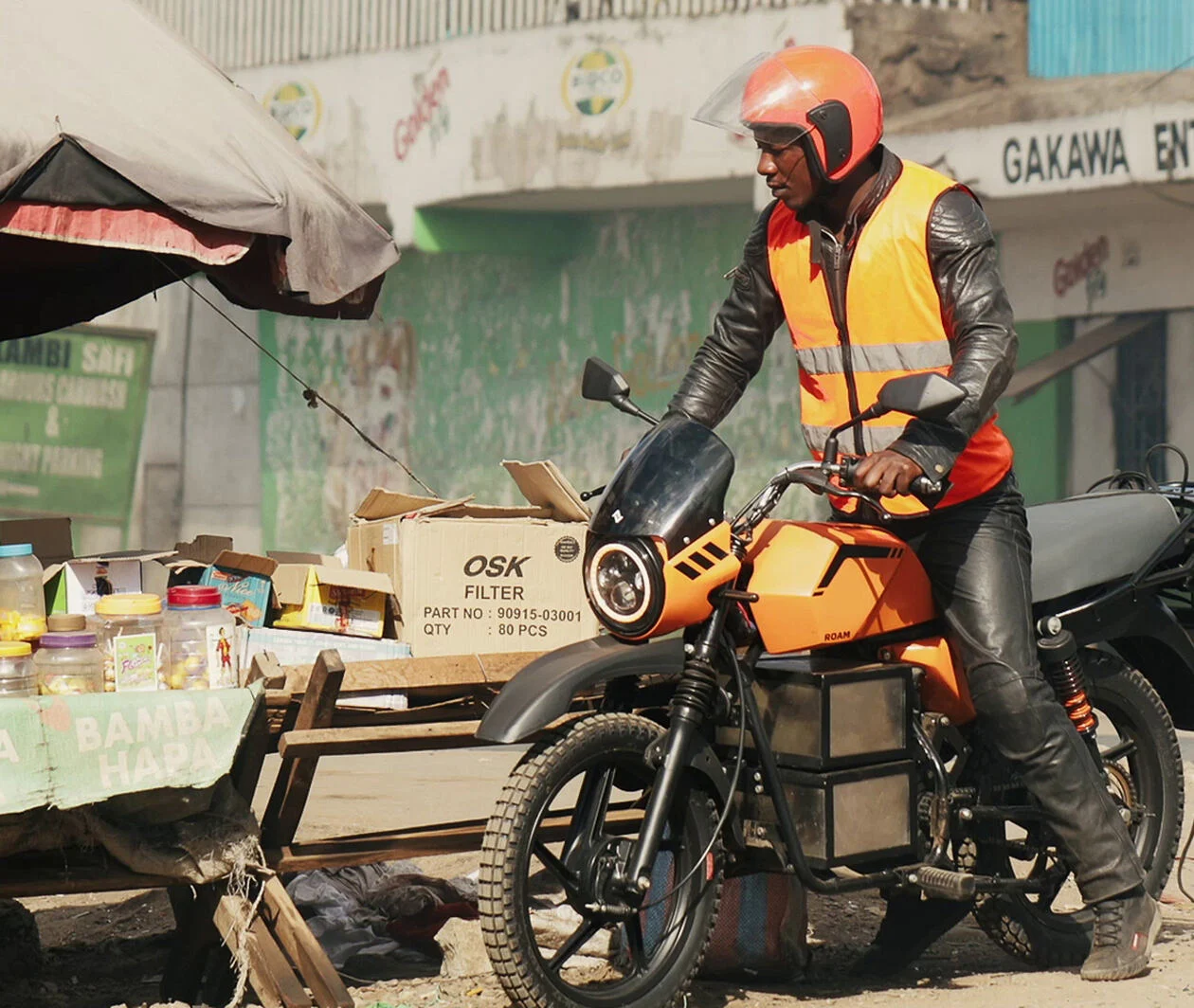 A man with helmet and west standing on an orange electric motorbike on a african market. 