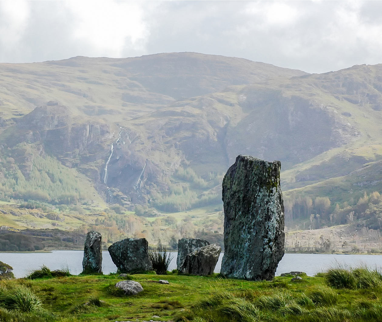 Standing stones on a grass mound set against a backdrop of mountains and a lake.