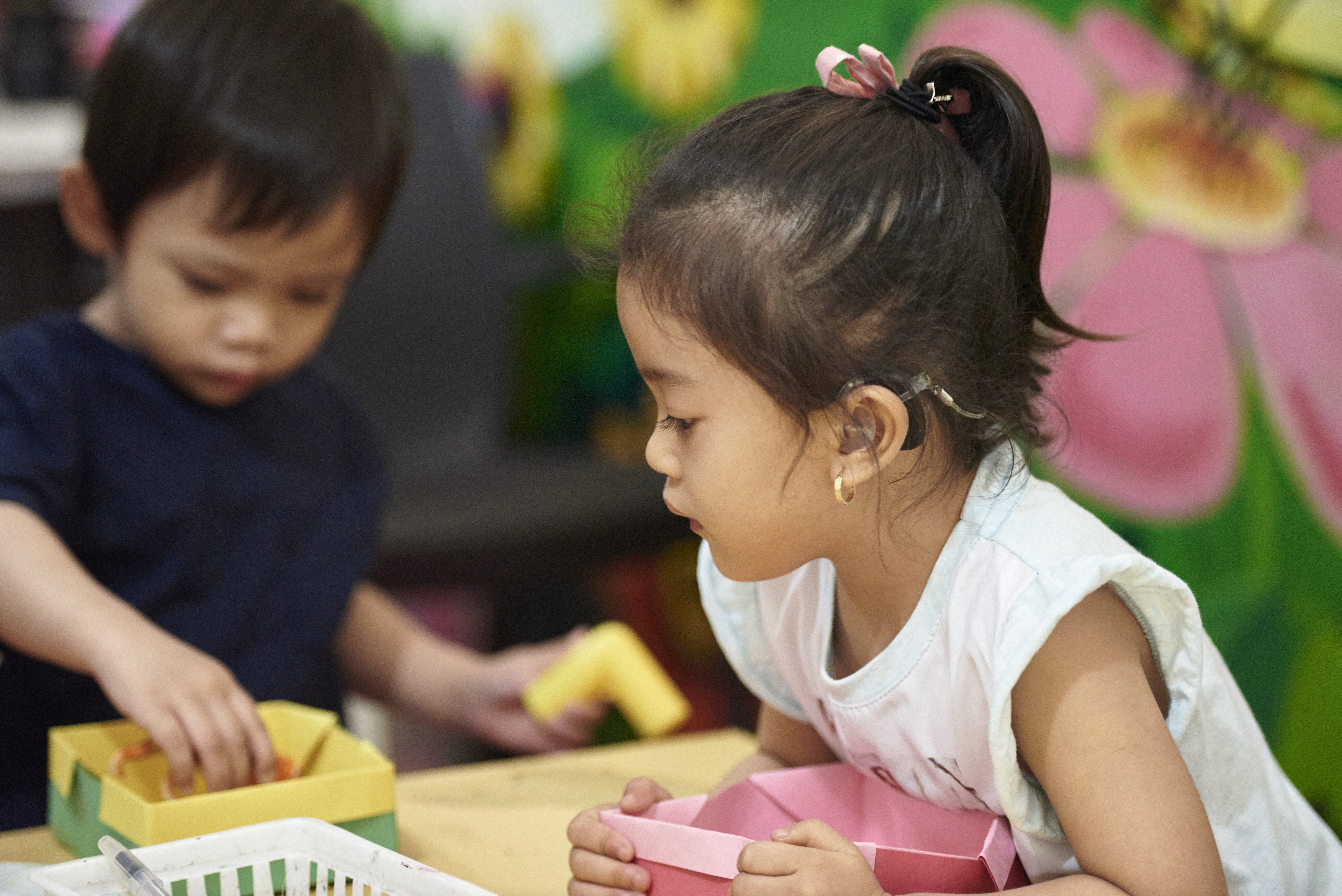 A young girl wearing a hearing aid is clutching a pink cardboard box in a school.