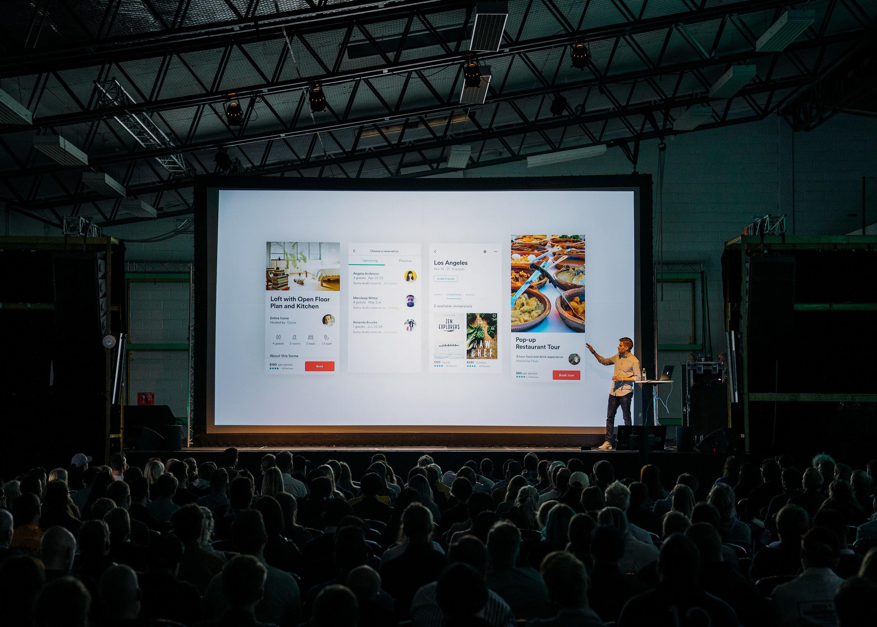 Person talking whilst standing in front of a large screen in front of a large audience