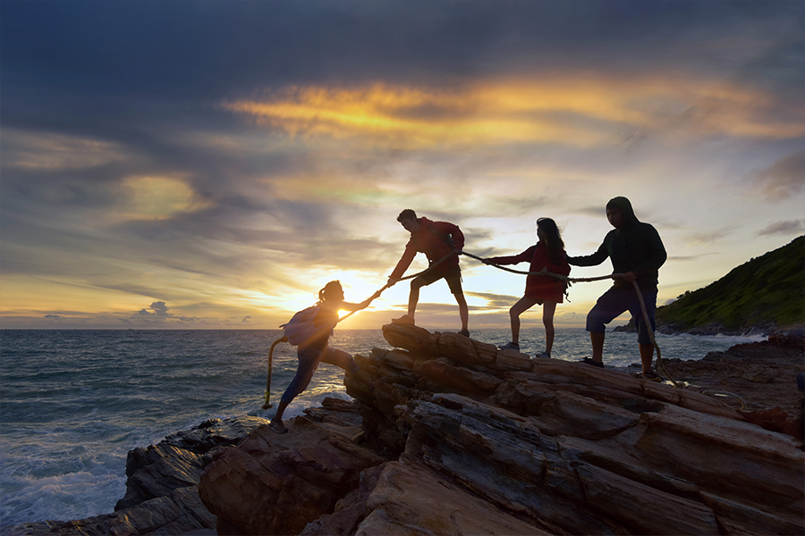 Three peopling standing on a cliff, using a rope to help a fourth person climb up. In the background there is a beautiful sunset over the ocean.