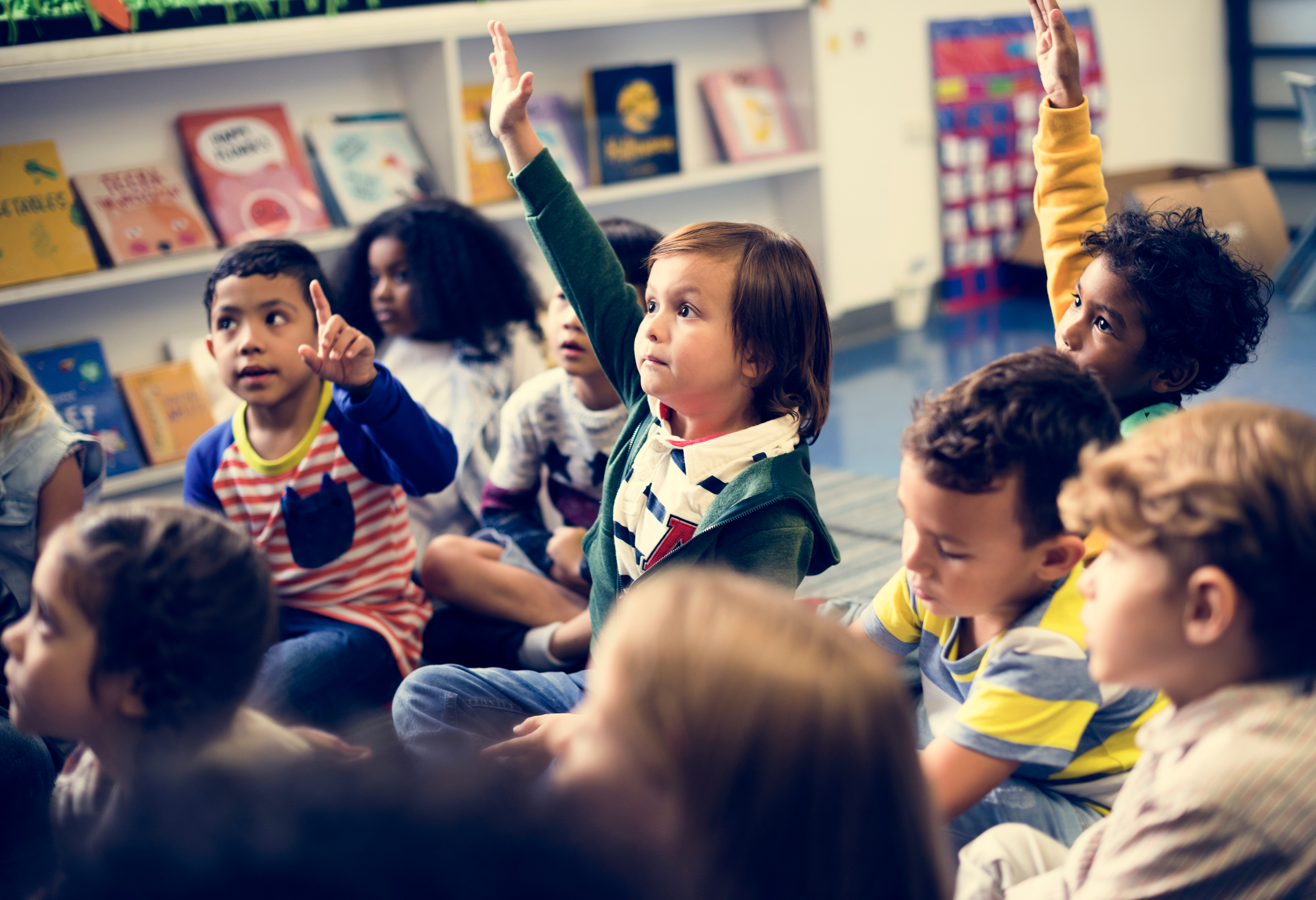 Group of children sitting on the floor in a classroom setting raising their hands