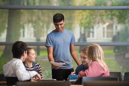 The image shows a group of undergraduate students seated round a table in a lecture building at the University of Nottingham while another smiling student stands talking to them.