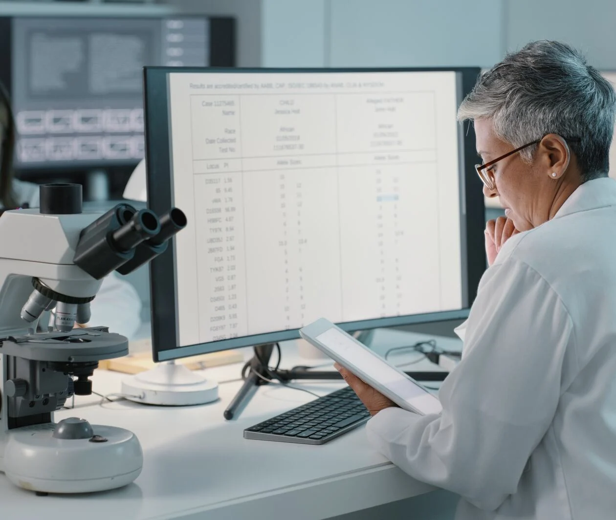 Woman wearing a lab coat and sitting in a biology laboratory reads off an iPad in front of a PC screen full of genomics data.