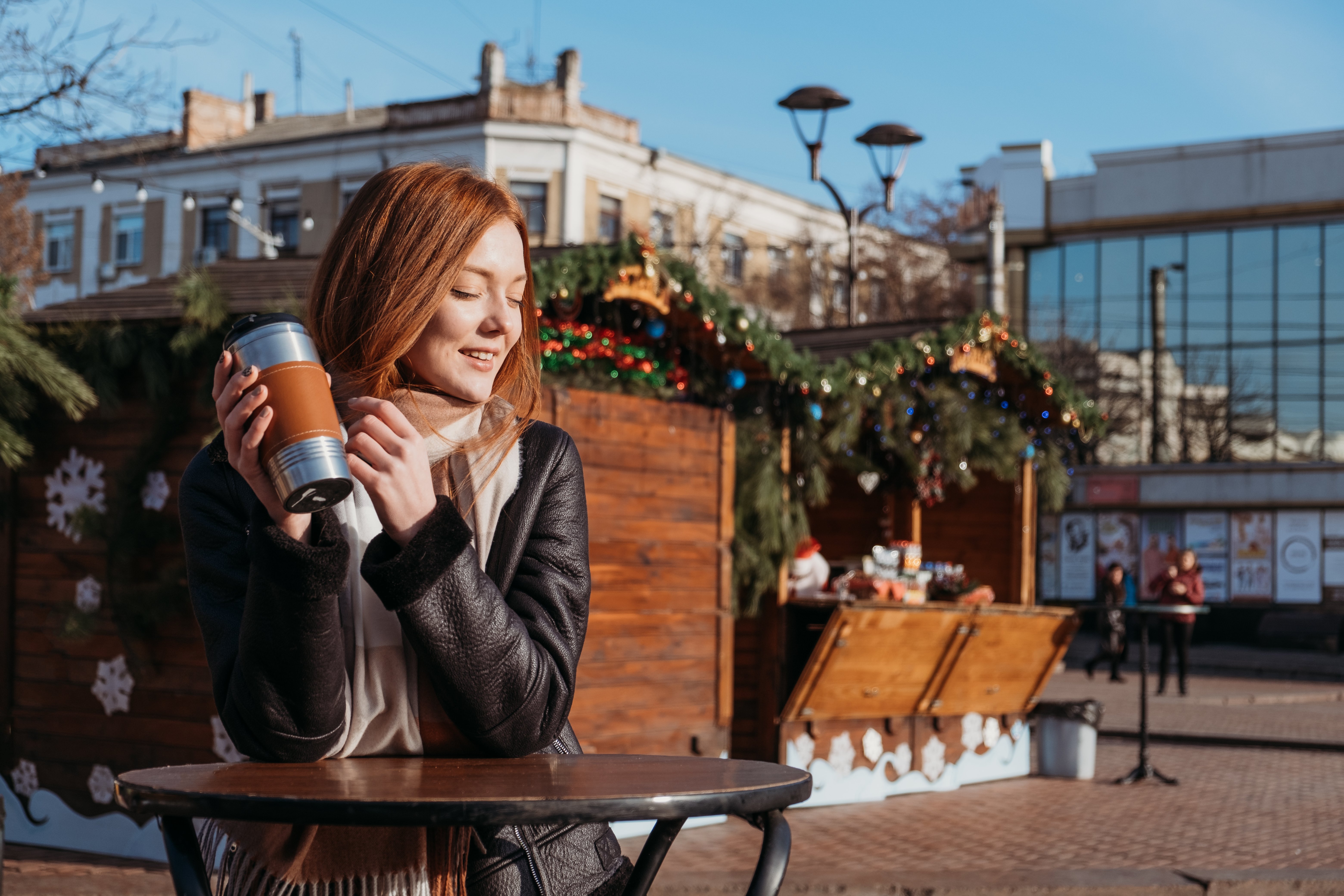 Woman standing in the winter sun at a table near an outdoor coffee stand. She has a reusable cup in her hand.