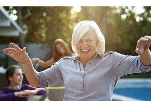 A playful image of an older woman, a teenager and young girl having fun hula hooping in a back garden next to a swimming pool.