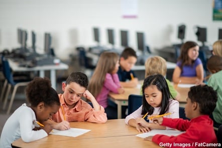 A group of four young pupils sitting at a big desk, each with a pencil in their hand. They are all working on a written task. Another group of six pupils can be seen out-of-focus in the background.