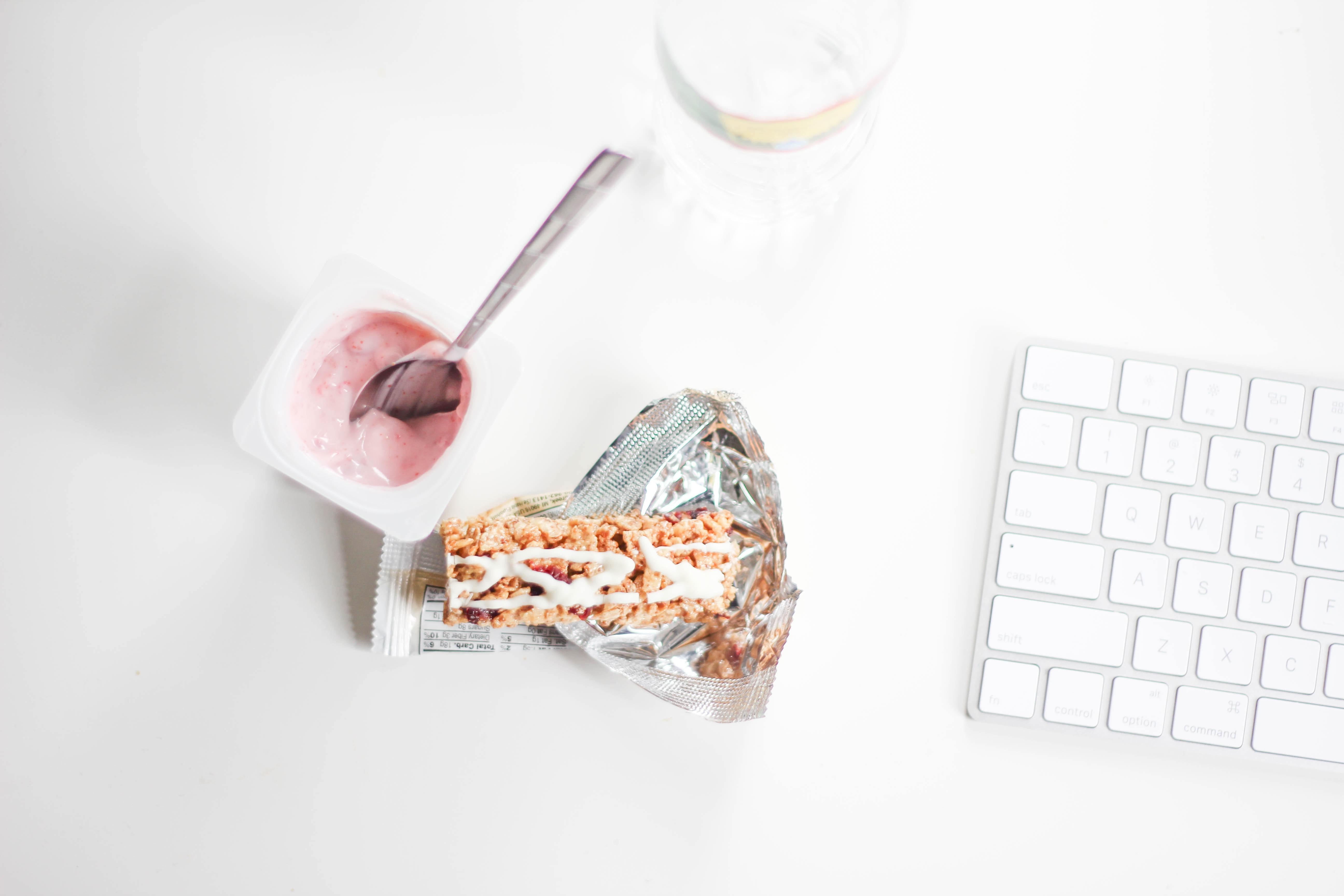 Photograph of an opened yoghurt next to an unwrapped ceral bar by a computer keyboard