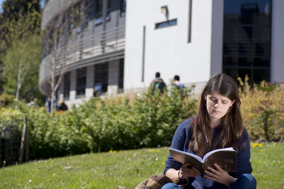 The image shows a student sat outside the Mathematical Science building on University Park Campus in Nottingham studying from a textbook.