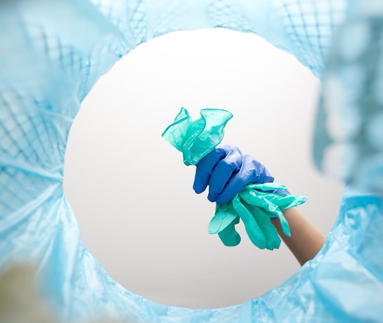 Gloved hand discarding used disposable gloves into a blue-lined trash bin, seen from inside the bin.