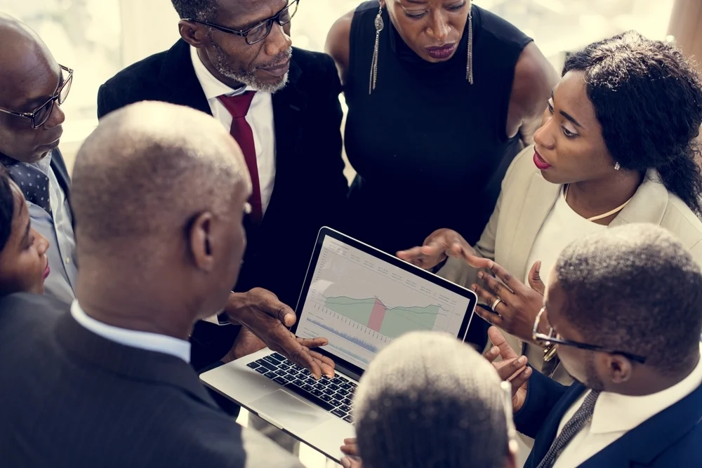 A group of people in business attire talking while looking at a chart on a laptop screen.