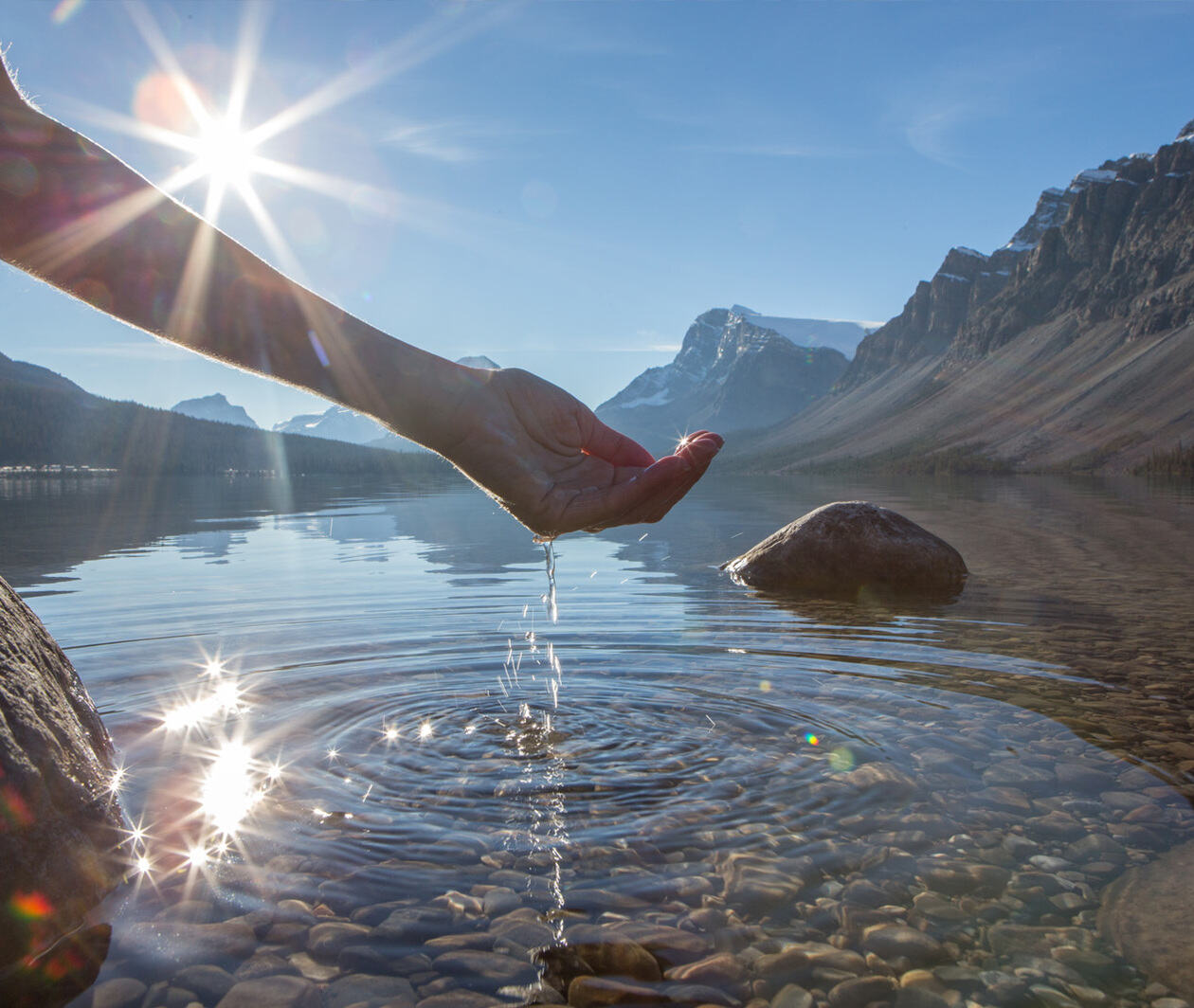 A wide shot if a human hand cupped to catch the fresh water from the lake, sunlight from sunset passing through the transparence of the water surrounded by ice mountains.