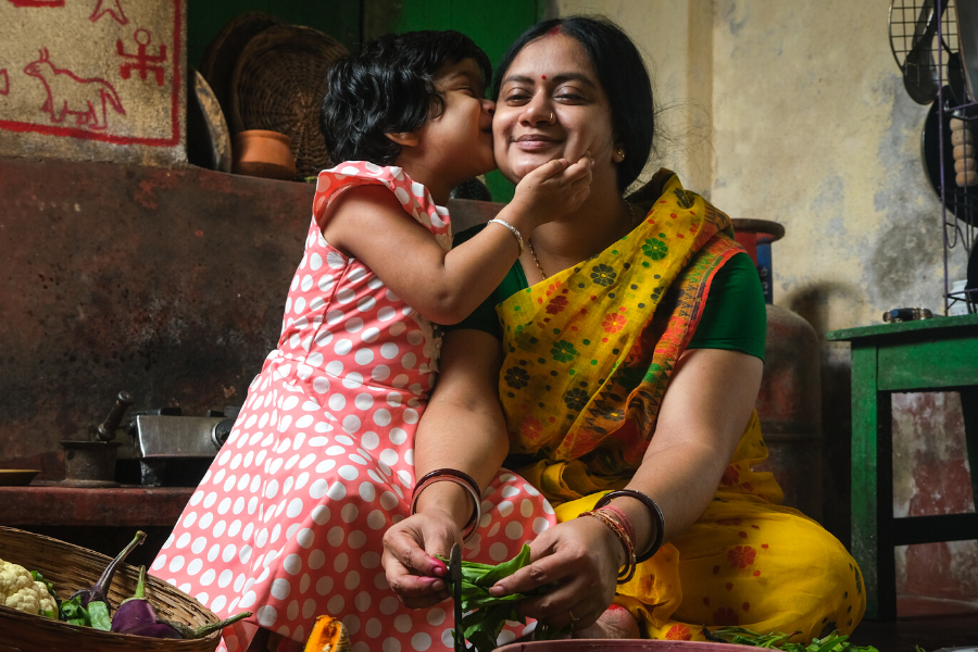 Child with dark hair and pink dress with white spots kissing woman on cheek who is seated on floor wearing yellow sari with flowers and preparing vegetables.