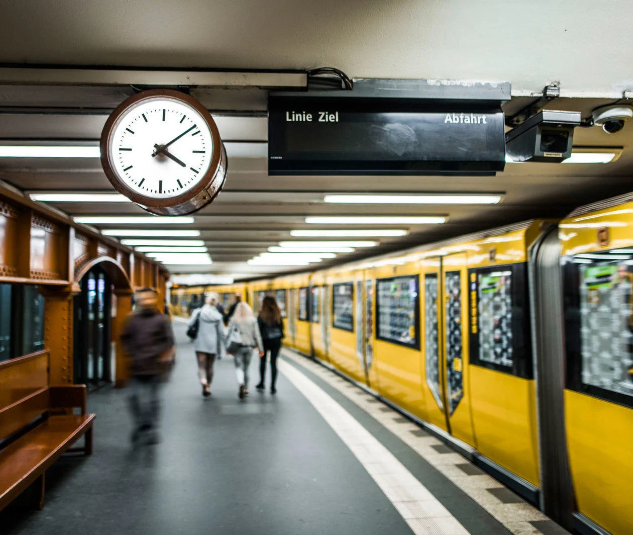 A station platform on a German underground railway, with a clock and train indicator board and several passengers walking beside a bright yellow train.