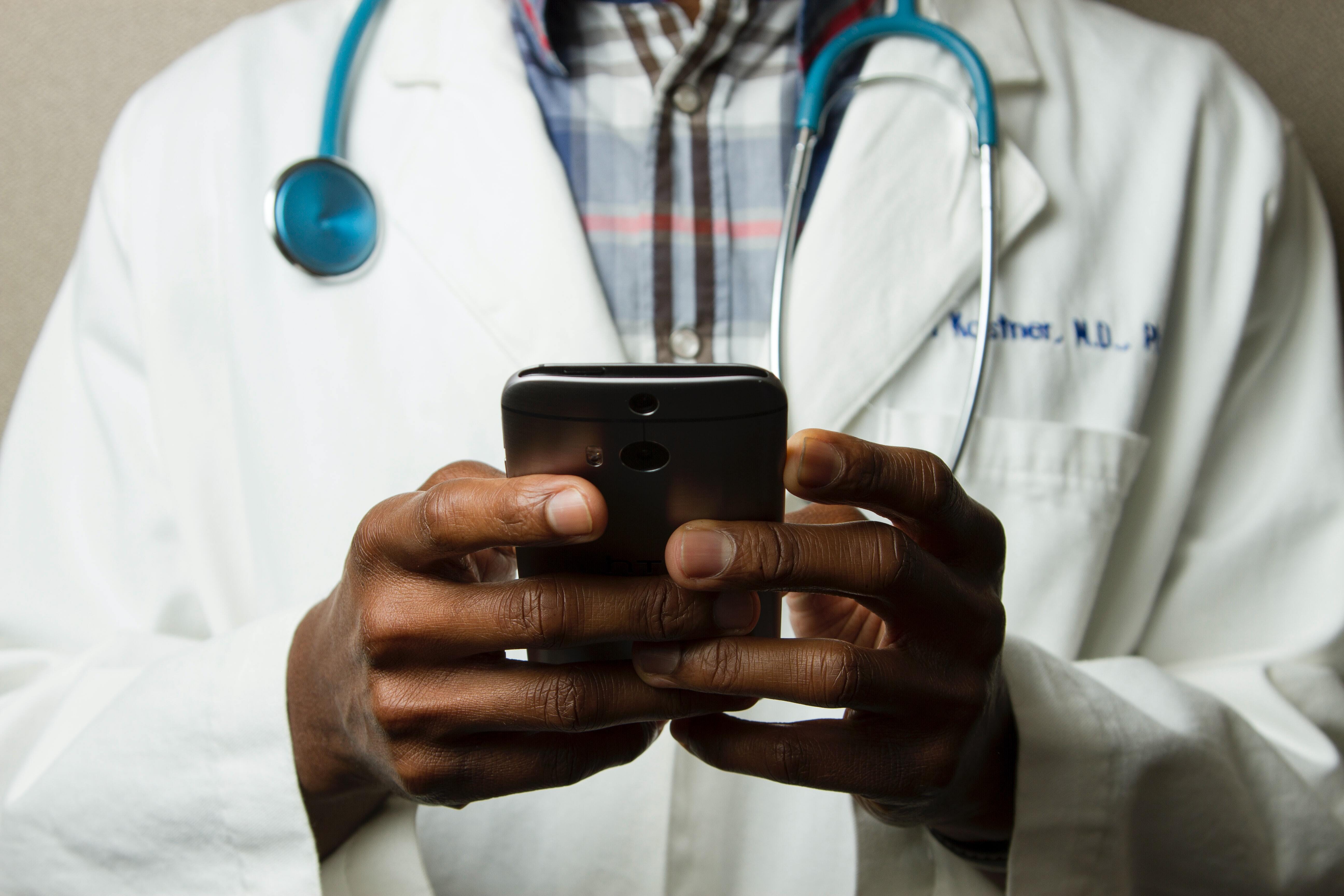 African American doctor in the US shown holding a smartphone. We see a close up of his hands and that he is dressed in a doctor's gown and has a stethoscope around his neck.