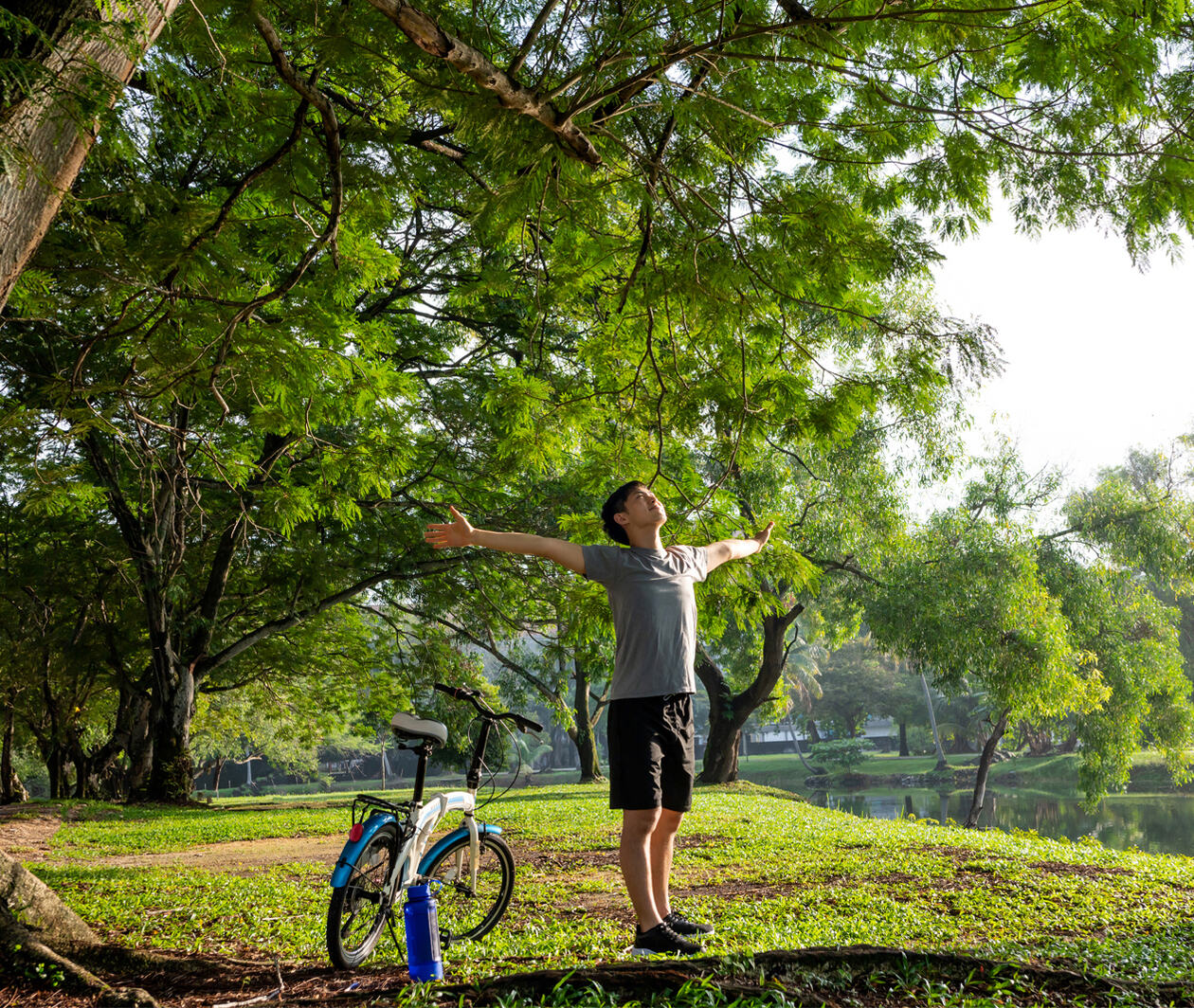 Young man breathing fresh air stretching arms in a park with big trees and a lake in the background and a bike by his side.