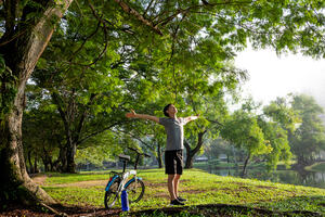 Young man breathing fresh air stretching arms in a park with big trees and a lake in the background and a bike by his side.