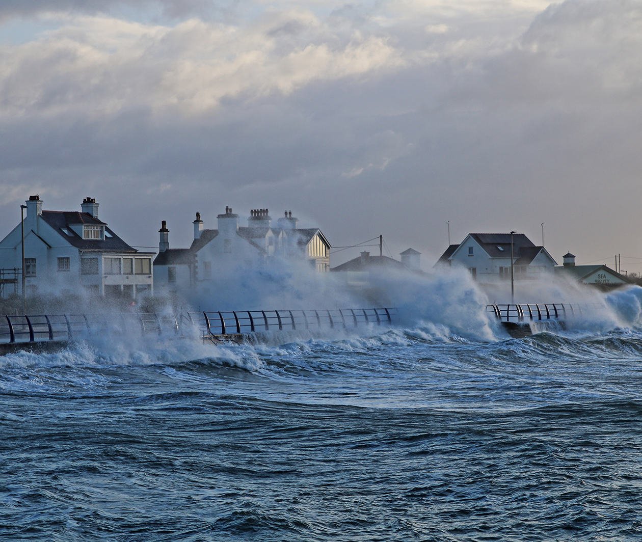 The picture shows a stormy sea, with waves whipping up over the sea wall onto the promenade.  