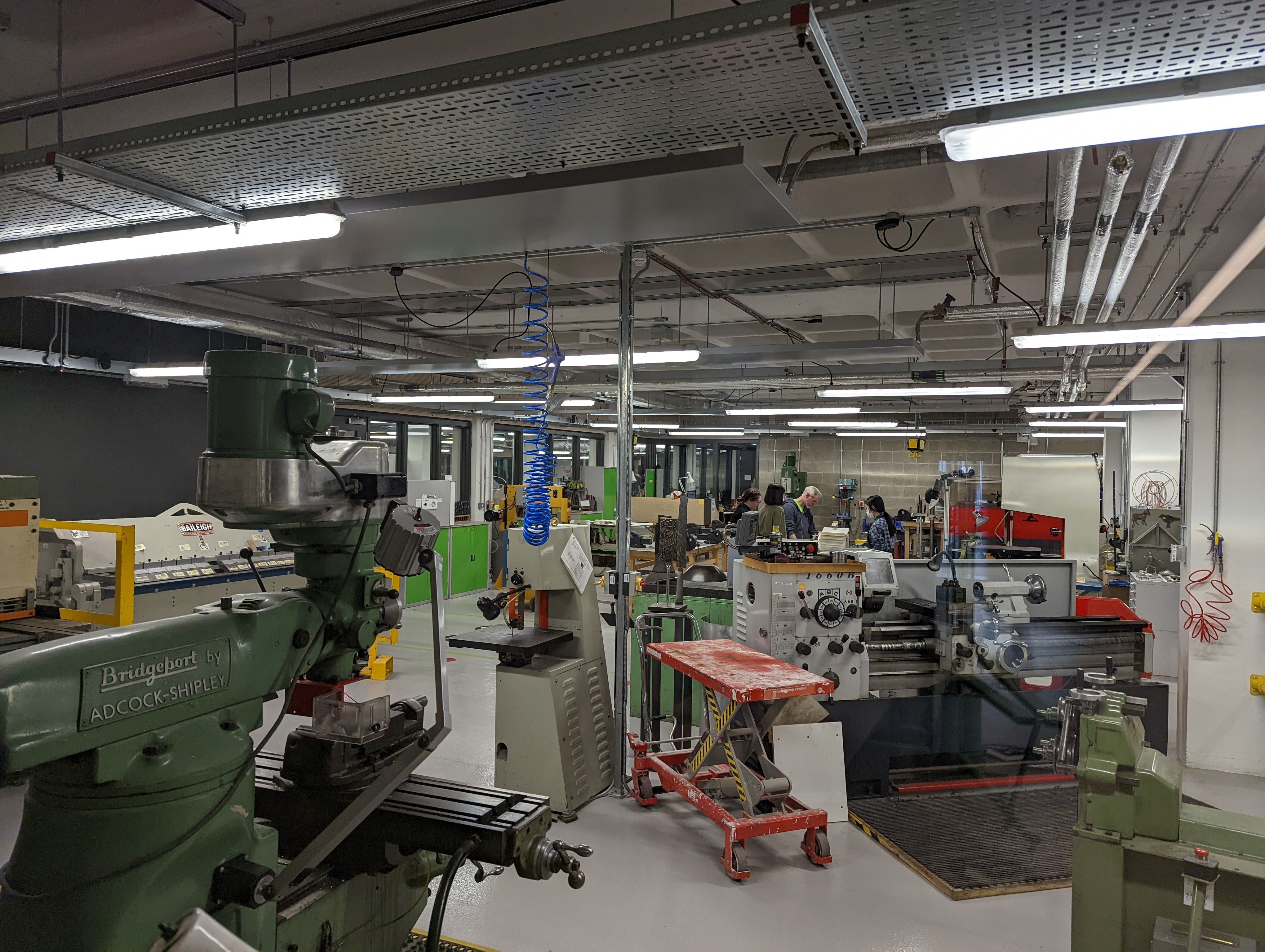 Kingston's metal workshop room. A large room with ceiling strip lights filled with large machinery.