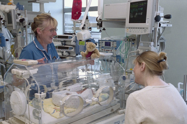 An image of a special care baby unit, showing two adults next to an incubator with a small baby. A teddy bear is on top of the incubator.