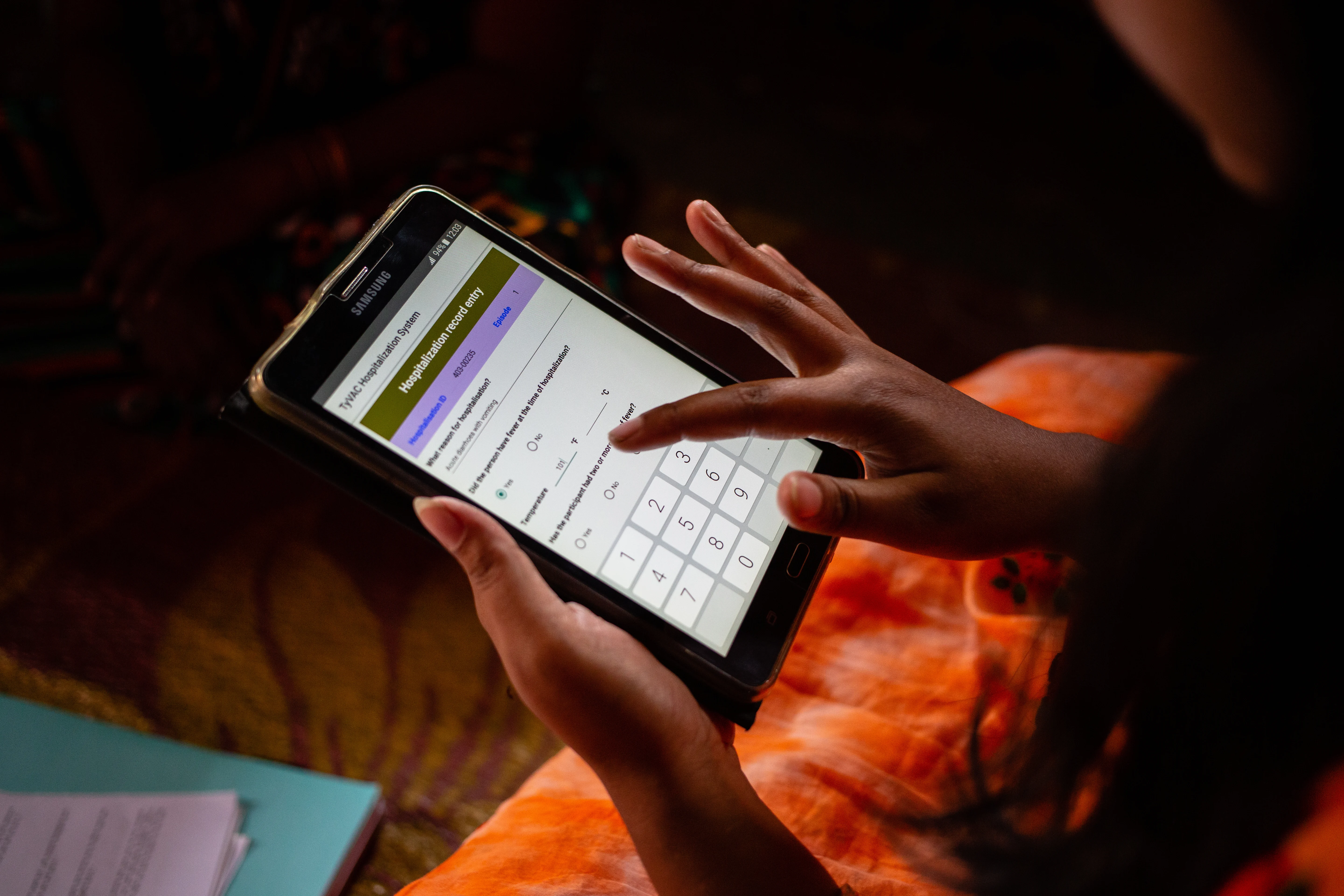 A community health worker is holding a tablet. The tablet is showing hospitalisation record entry on top, with two questions in the middle and a dial pad at the bottom.