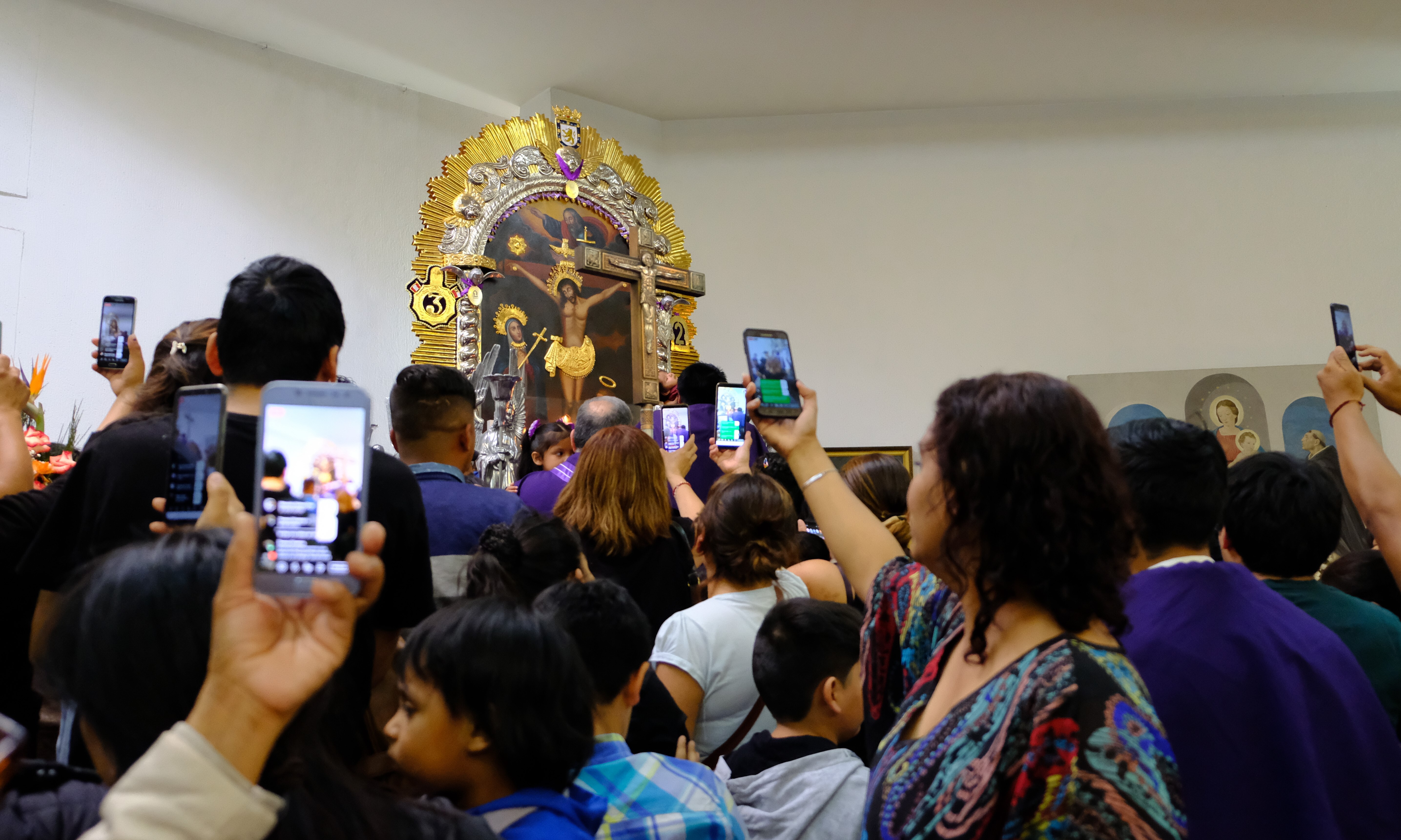 Interior of a church with a painting of the Christ, surrounded by people aiming their smartphones at the image. These are Peruvian migrants honoring in Chile the Lord of Miracles, the most revered Peruvian religious icon.