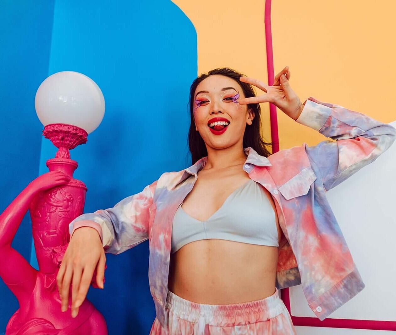 A woman smiling with red lipstick and colourful purple eye make up posing in front of a red, blue and yellow wall. 