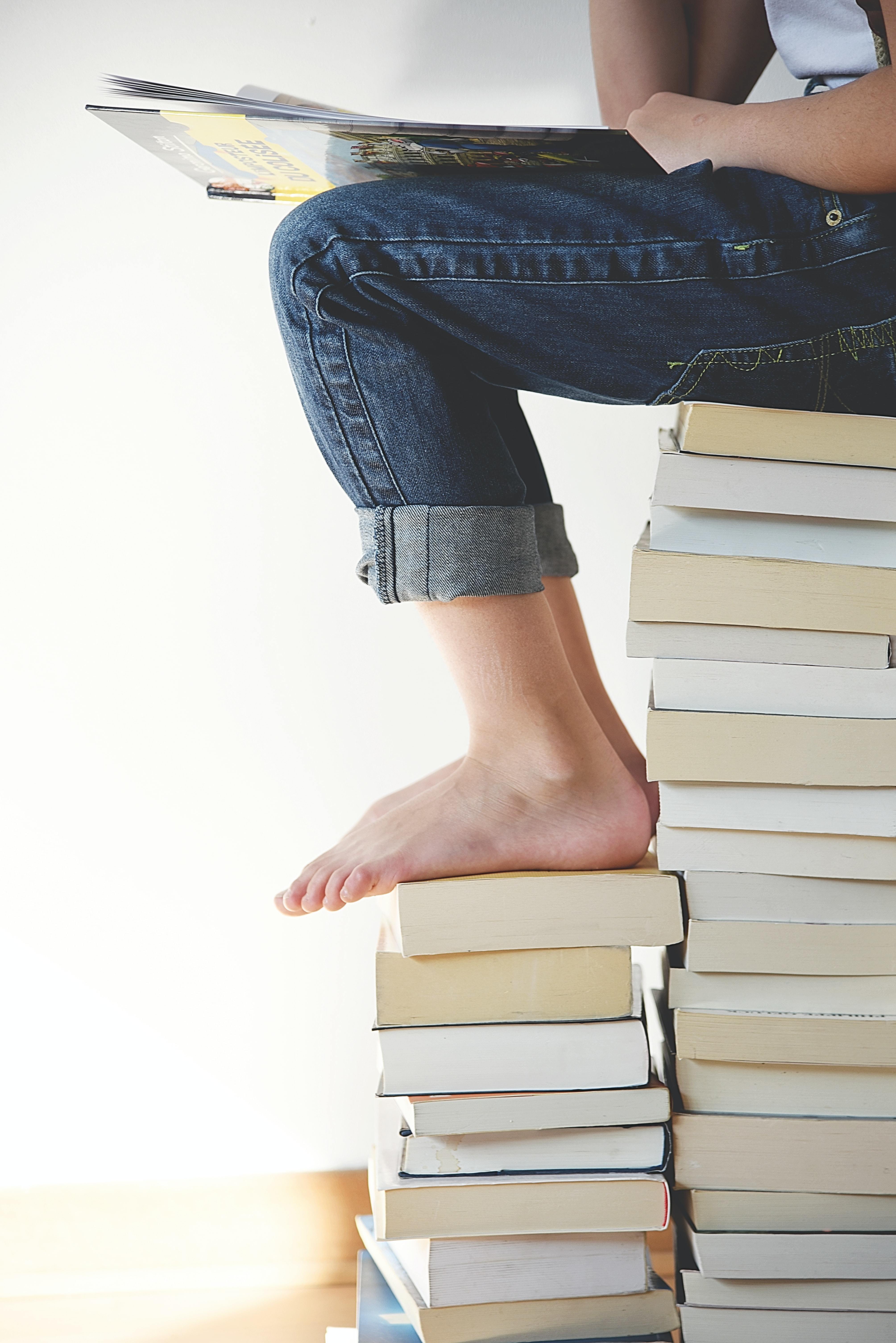 Lower legs of a young person, in jeans, sat atop a stack of books - viewed in profile.