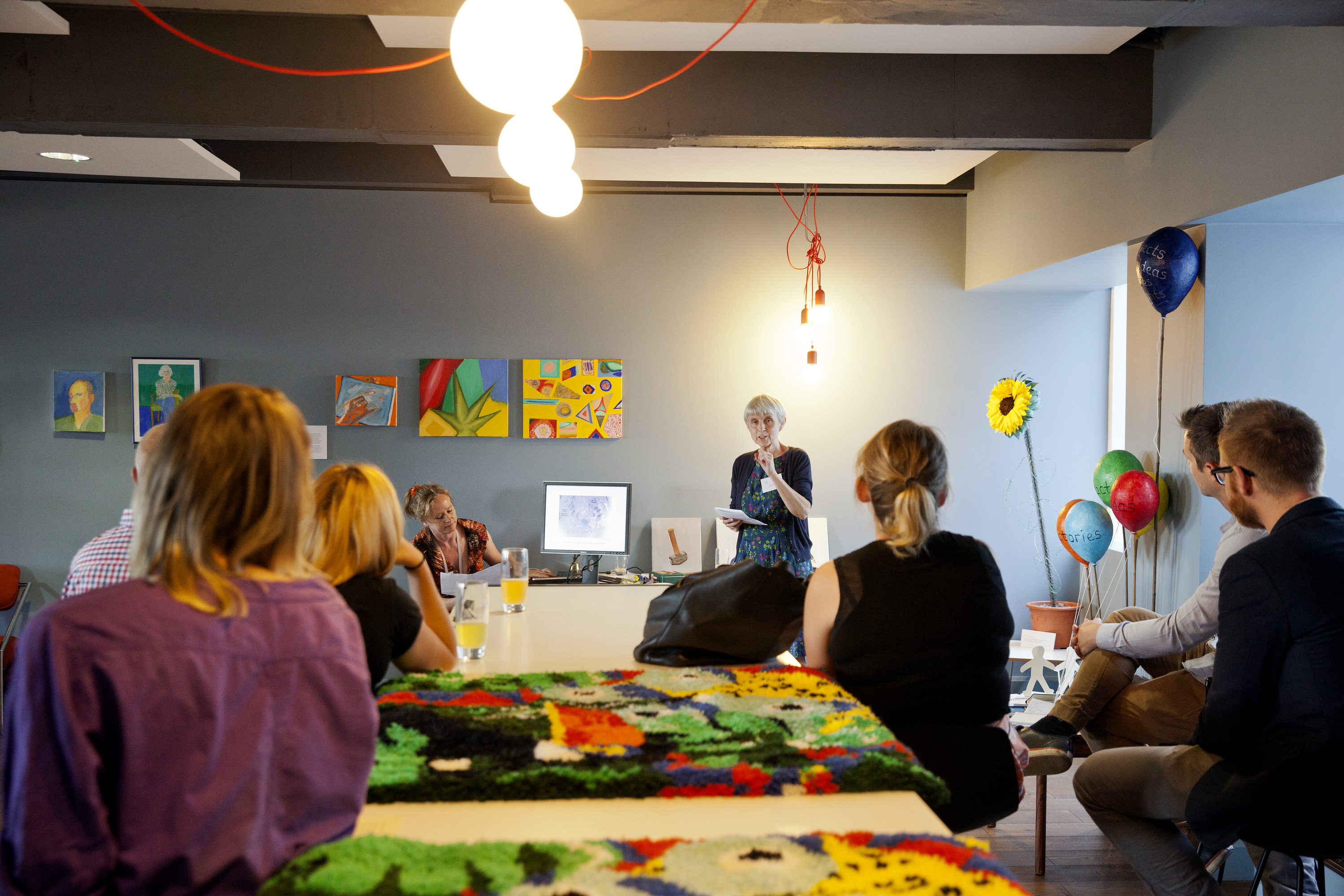 Photograph of a group of people sitting among various artworks, listening to a speaker