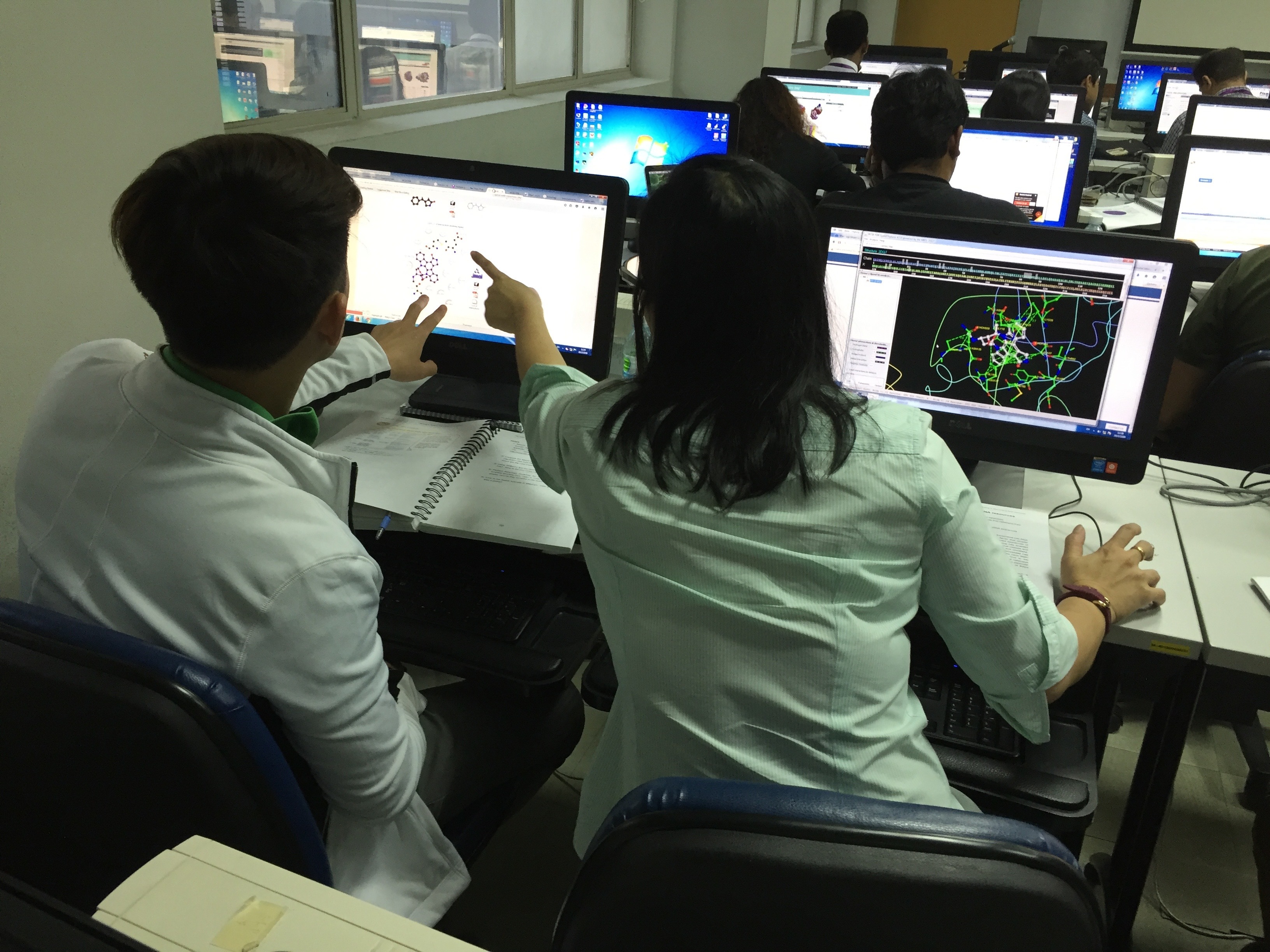 Back view of two seated learners (on the right a man, on the left a woman) engaging in discussion; with one pointing at the other's screen as they talk with each other about the analyses displayed on their monitors.  The context a computer room