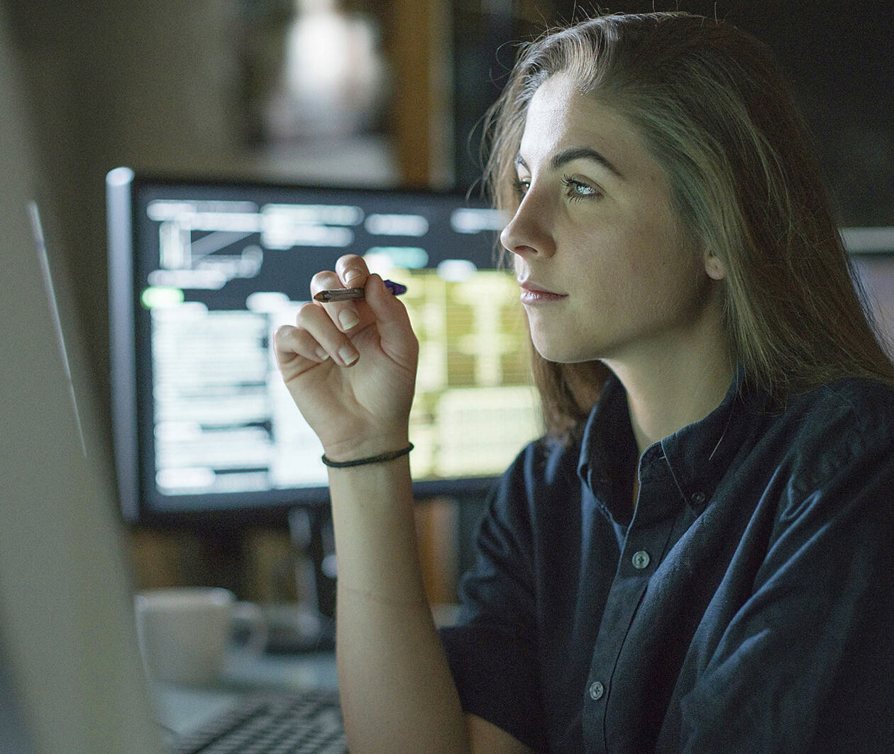 A woman with a pen in her hand looking at a screen. There's a screen in the background too.