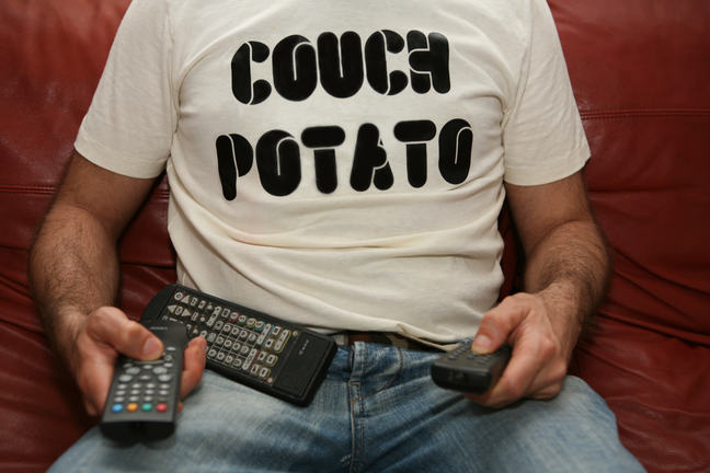 A man wearing a t-shirt reading 'couch potato' sits on a sofa holding remote controls