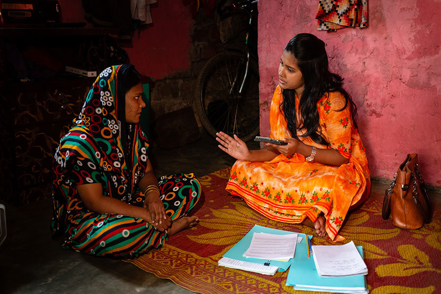 Two women are sitting down on a mat. The woman on the right, is holding a tablet and is conveying information to woman on the left, who is listening intently. In front of them are an assortment of papers and notebooks with pencils.