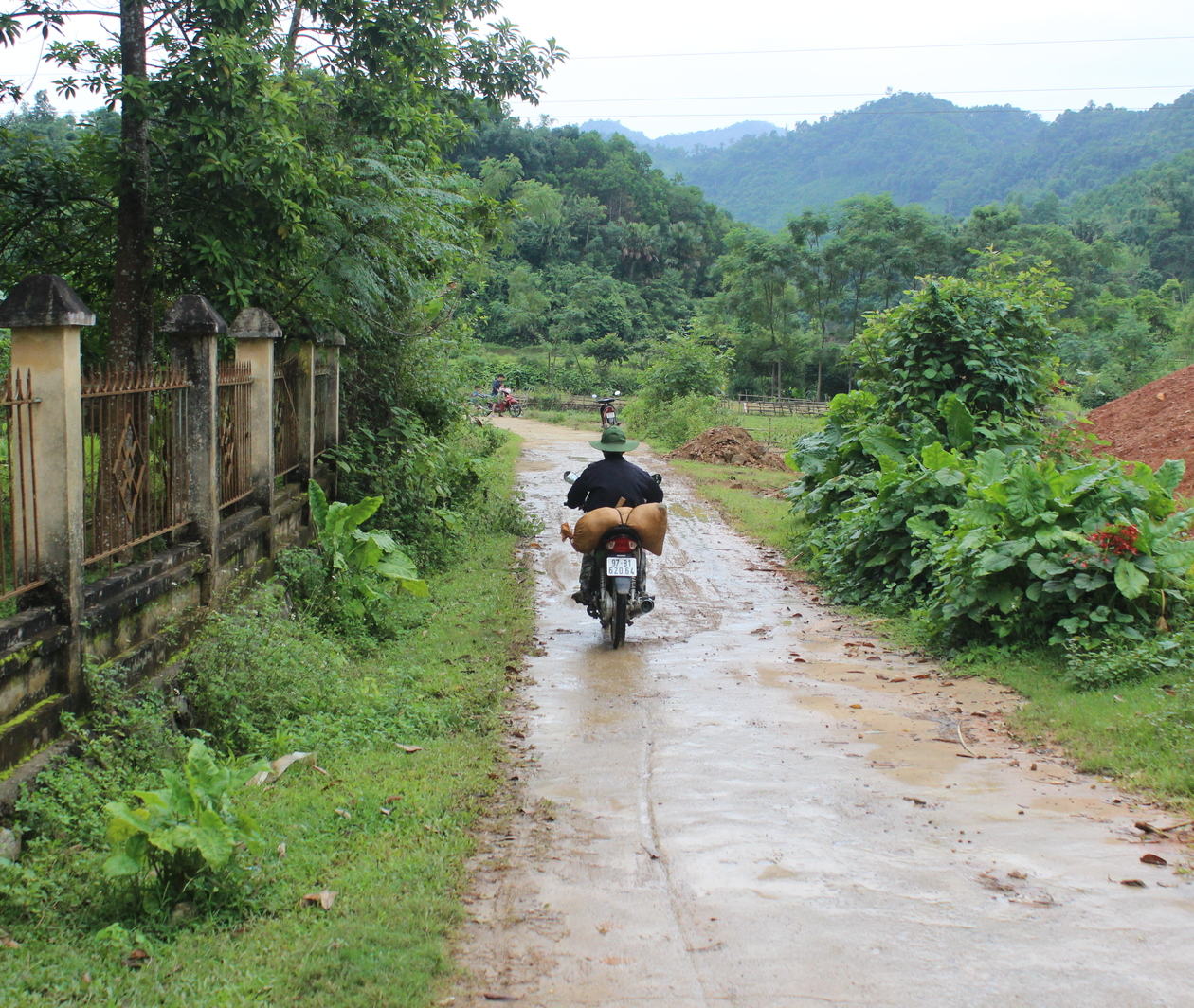 A narrow rural road in Vietnam with mud caked surface and puddles. A single motorbike with a sack tied to the back leaves a tyre track