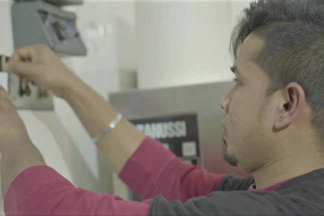 A young man with short spiky dark hair and wearing a red and black shirt is pinning a piece of paper to a noticeboard in a commercial kitchen.