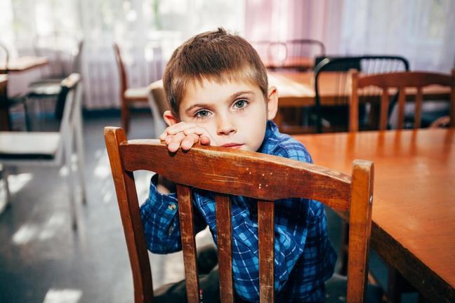 A young boy sits facing the back of a wooden chair with his hands resting on the top and his cheek resting on his hands. He is wearing a blue shirt. The room he is in has many tables and chairs, all mismatched.