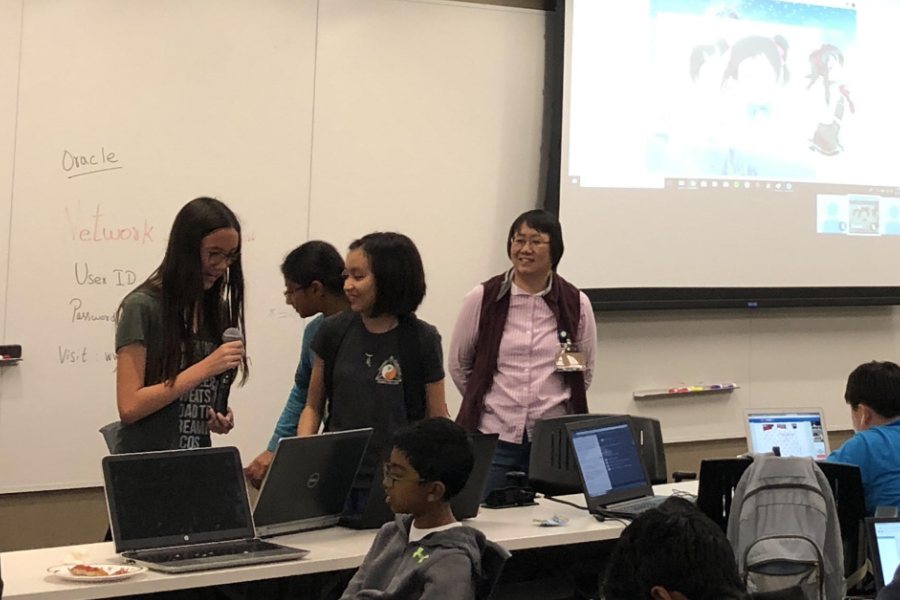 Two young people stand in front of a projector screen, a laptop in front of them, presenting to the group. One holds a microphone.