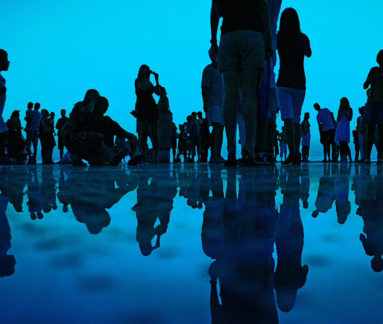 Group of people standing by the ocean, their reflections mirrored in the water below