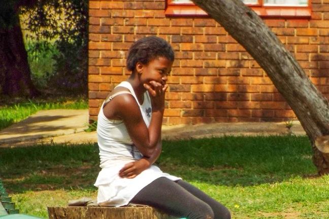 Teenage girl sitting on log in front yard with chin in her hand.