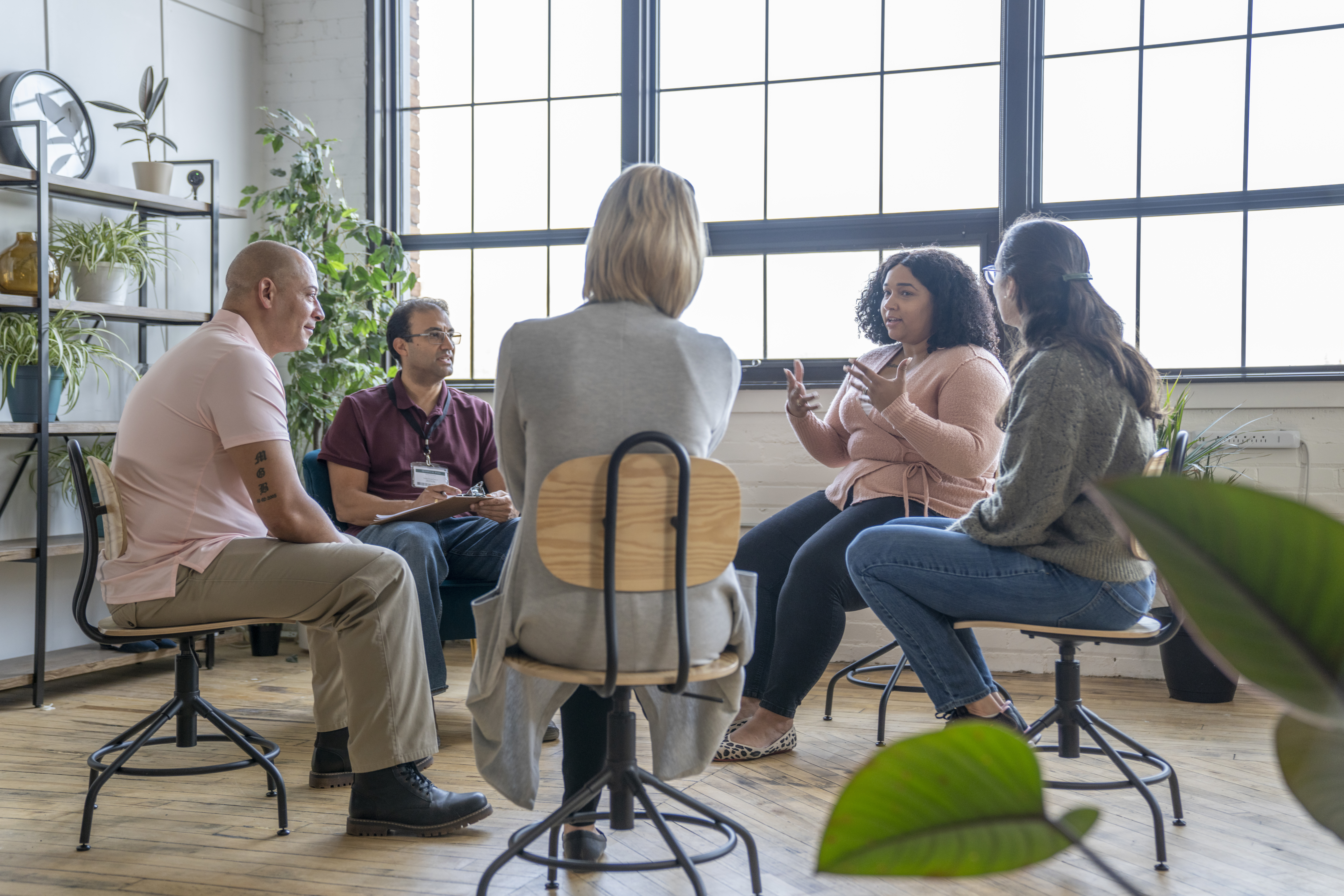 Multiethnic group of adults gather to discuss their mental health, addictions, and struggles in a group setting with a professional leading the discussion.