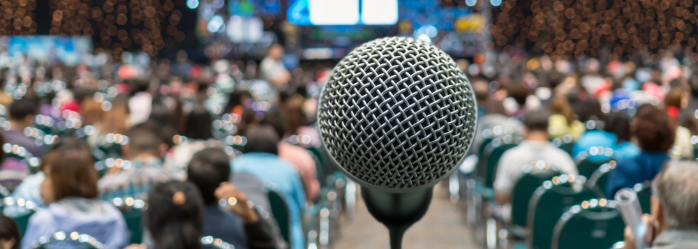 A microphone is shown in front of a blurred conference hall in the background with speakers on the stage and the audience in the background.