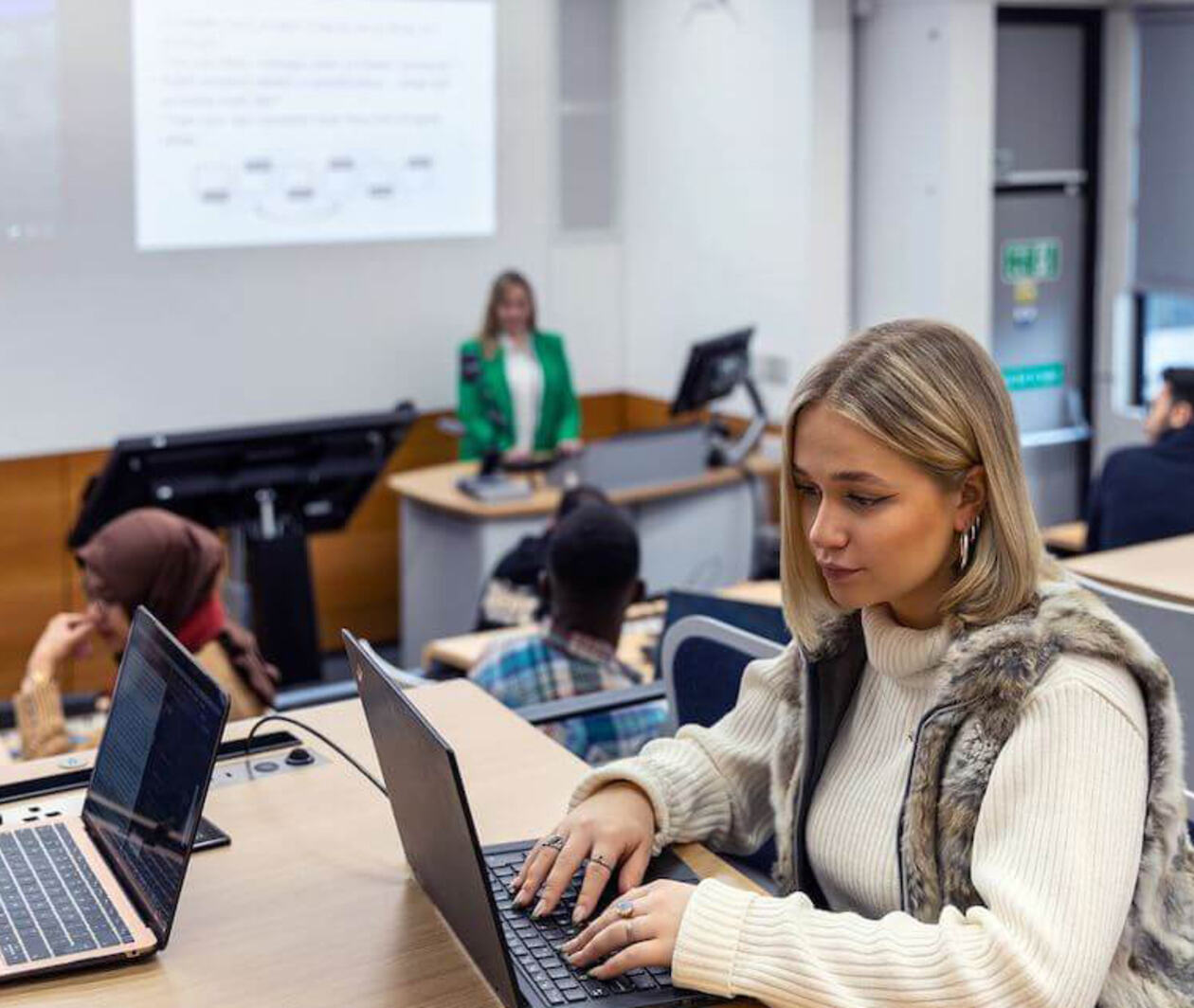 Students using laptops in a university lecture hall while a lecturer presents at the front of the room