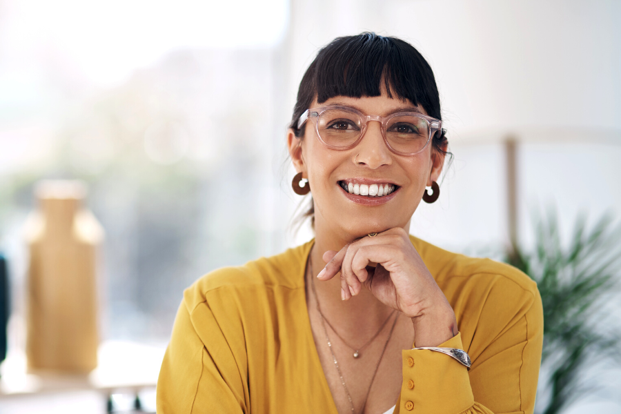 Woman in mustard blouse, pink glasses and nose ring smiles at camera.